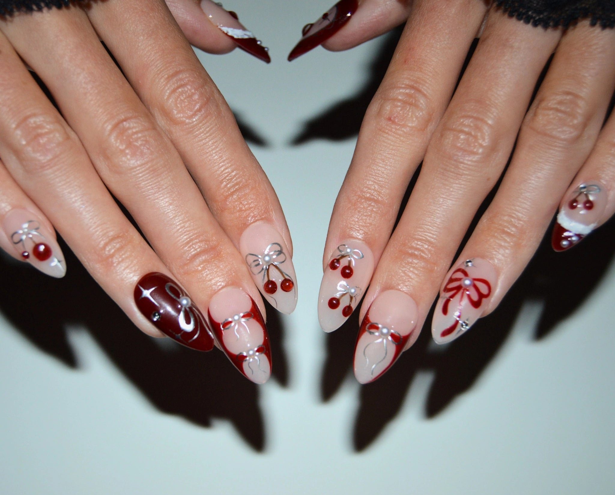 Press-on nails Cherry Christmas faits main en gel et acrylique, design nude et rouge avec cerises 3D, nœuds métalliques et finitions festives.
